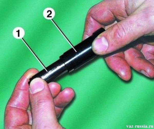 В руках человек держит натяжитель, а стрелкой один указан плунжерная часть его, а стрелкой два указан сам корпус натяжителя